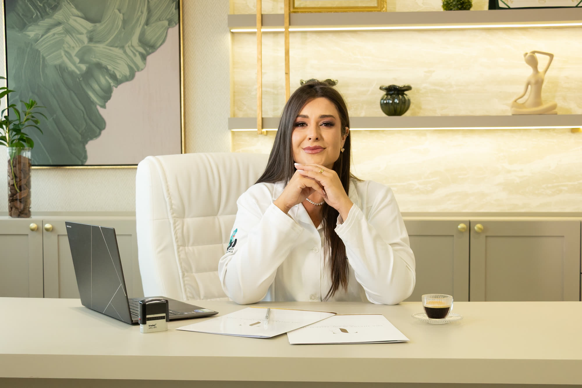 Dra. Thaynã Alane no consultório, apoiada na mesa de atendimento, com prontuários e computador à frente.