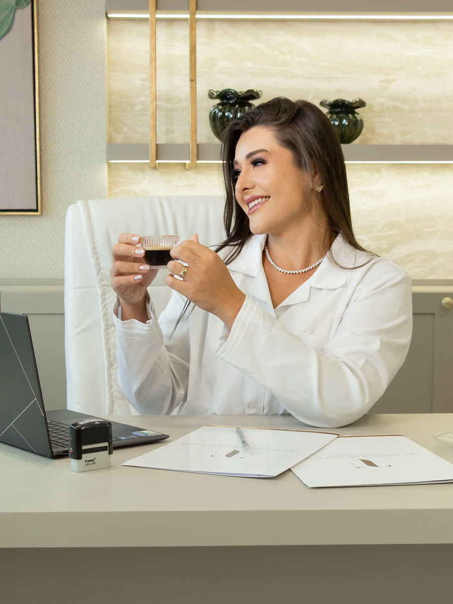 Dra. Thaynã Alane sorrindo no consultório, com uma xícara de café sobre a mesa de atendimento.
